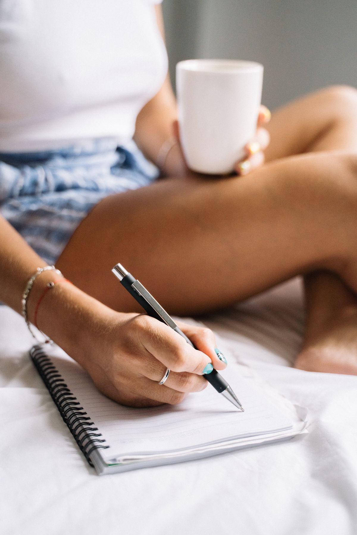 A woman sitting on a bed writing in a notebook with a mug nearby — keeping a sleep diary.