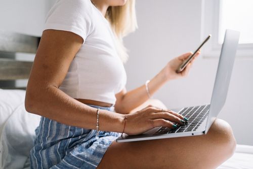 Free Woman multitasking with laptop and phone, working comfortably from home on a bed. Stock Photo
