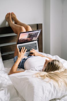 女人躺在床上，把腿搭在床头板上，用着笔记本电脑，室内环境很休闲