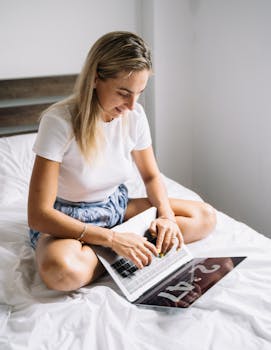 Young woman in casual wear working on her laptop while sitting on a bed indoors.