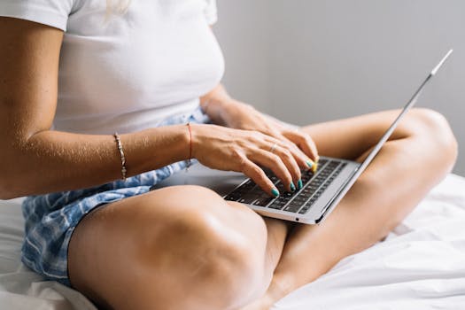 Unrecognizable woman typing on a laptop while sitting cross-legged on a bed, showcasing home relaxation and digital lifestyle.