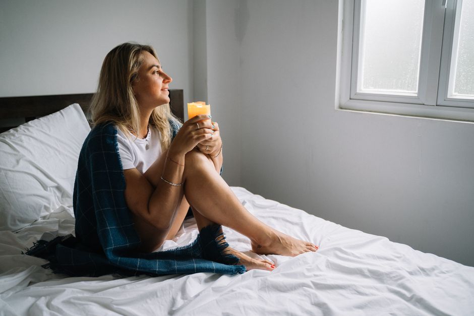 Woman sitting on bed with candle wrapped in a cozy blanket, enjoying morning tranquility.