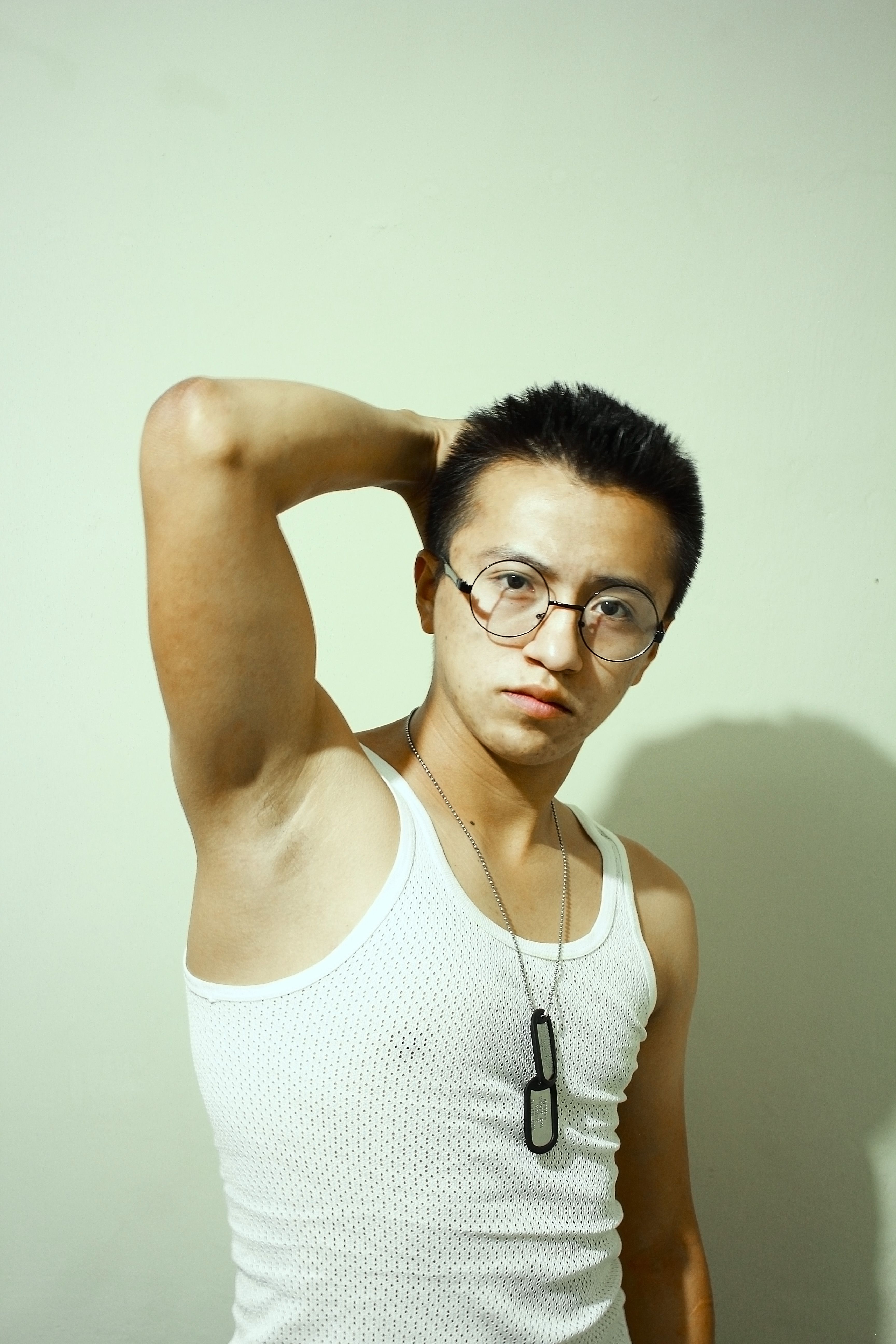Portrait of a young man in a white tank top and eyeglasses posing confidently against a neutral background.
