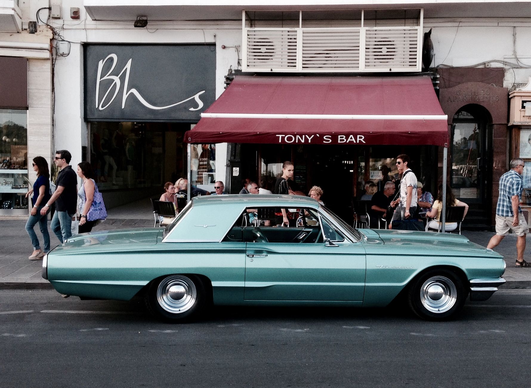 A retro car parked outside a bar with people on the sidewalk, capturing a lively street scene.
