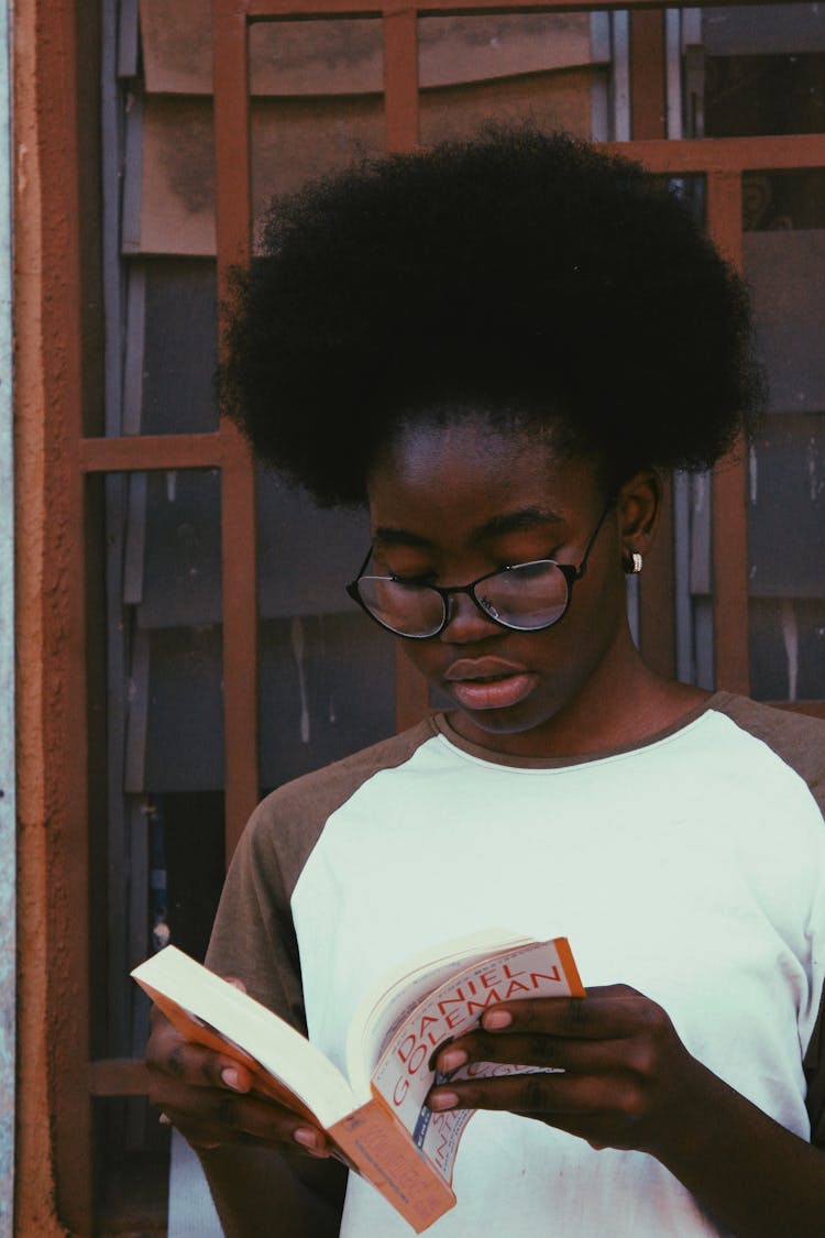 Black Student In Eyeglasses Reading Book Near Wall