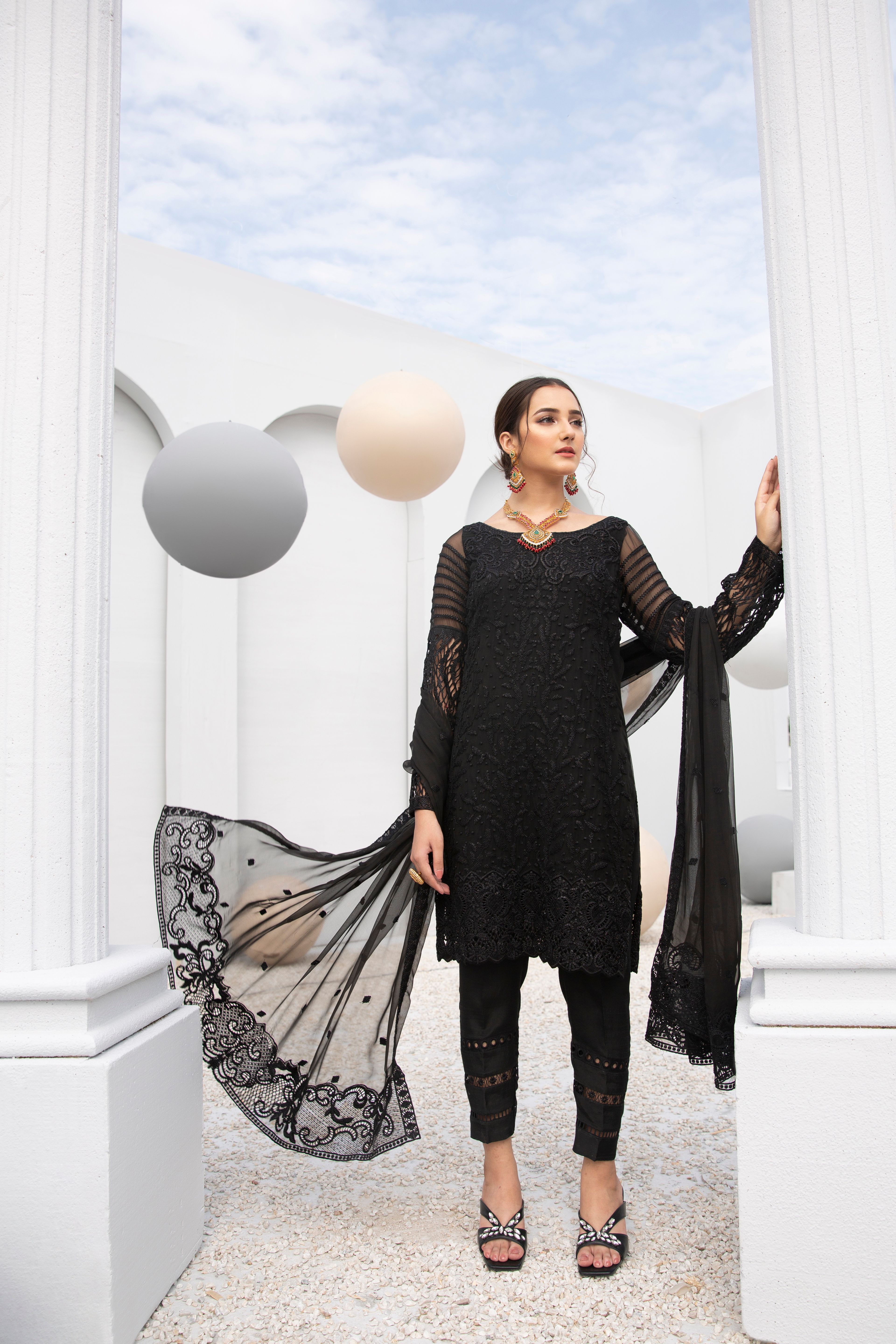 Woman posing in stylish black traditional attire amid elegant white pillars.