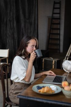 女人在室内一边用笔记本电脑工作，一边喝着咖啡，桌上还放着零食