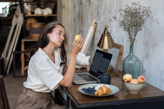 一位年轻女子在艺术工作室里，手拿桃子，坐在木桌旁使用笔记本电脑