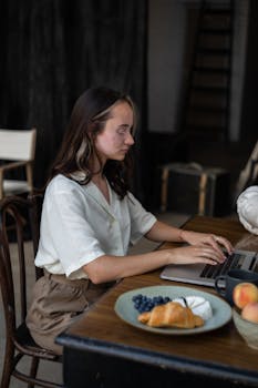 A woman concentrating on work at her laptop, sitting at a table with a breakfast meal.