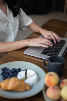 Woman working on a laptop with a healthy breakfast including cheese, fruits, and a croissant.