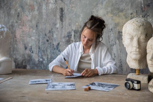 Female artist sketching at a table surrounded by sculptures and photos, exuding creativity.