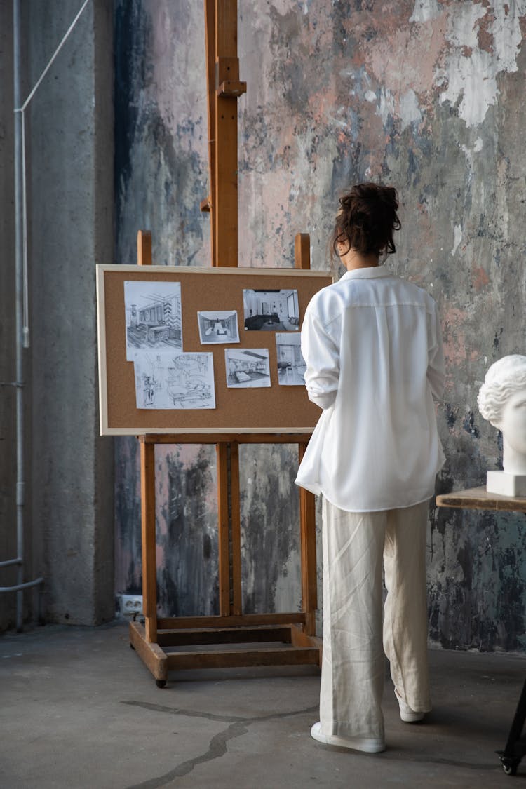 Woman In White Long Sleeves Shirt Looking At Drawings On A Corkboard