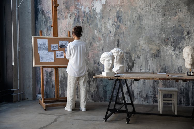 Woman In White Long Sleeves Shirt Looking At Drawings On A Corkboard