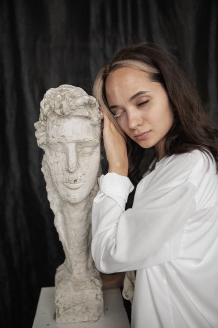 Woman in white shirt poses next to a gypsum sculpture with closed eyes, evoking a sense of serene art appreciation.