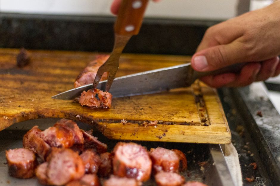 A hand slicing sausage on a wooden board with knife and fork.