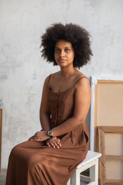 Elegant portrait of a woman with afro hair sitting on a wooden chair in a minimalist indoor setting.