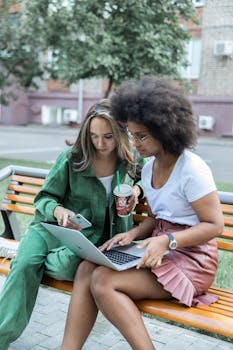两个女人坐在户外长凳上，一边喝咖啡一边用笔记本电脑合作