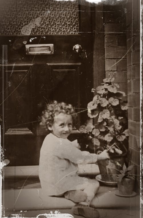 Grayscale Photo of Little Girl With Curly Hair Sitting On Doorstep Touching A Pot Plant