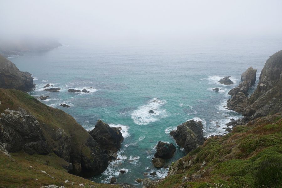 A serene foggy seascape with rugged cliffs and turquoise waves crashing against rocks.