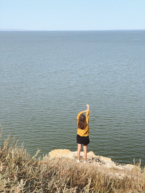 Back view of unrecognizable casual female traveler standing on rocky cliff with outstretched arms and admiring endless sea on sunny day