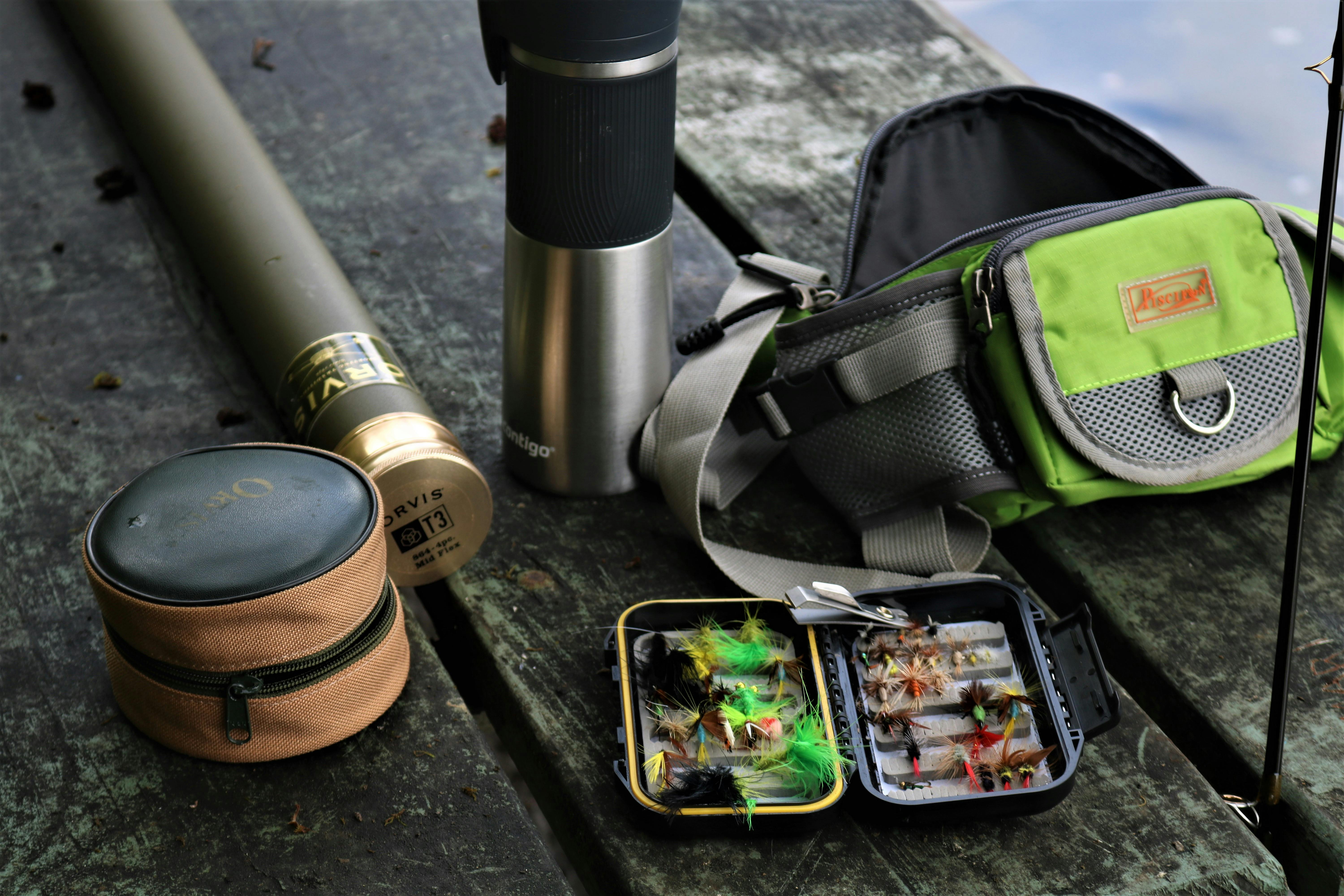 A detailed display of fishing equipment including fly box, backpack, and thermos, set outdoors.