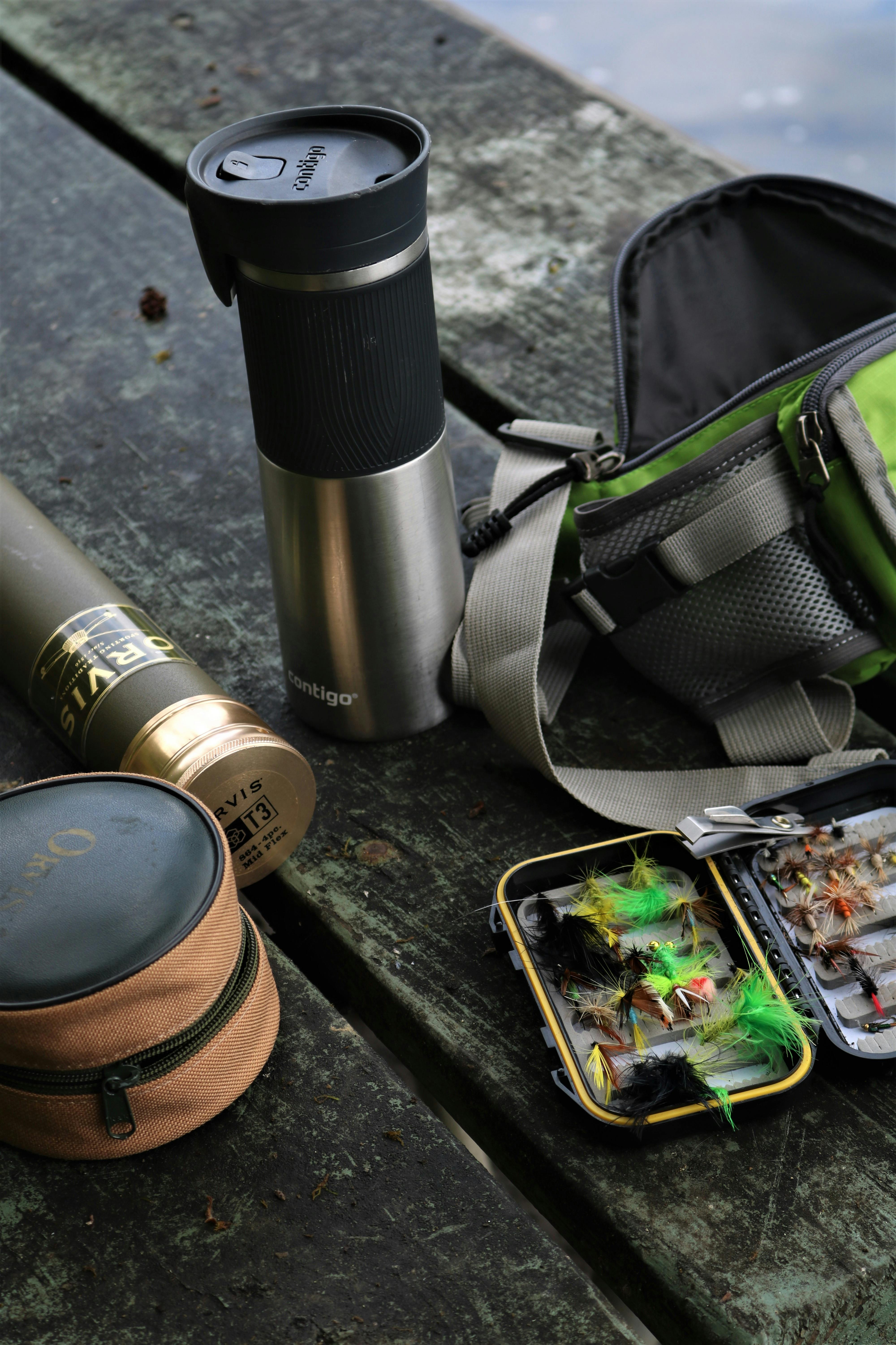 Fishing equipment including lures and a tumbler on a wooden picnic table outdoors.