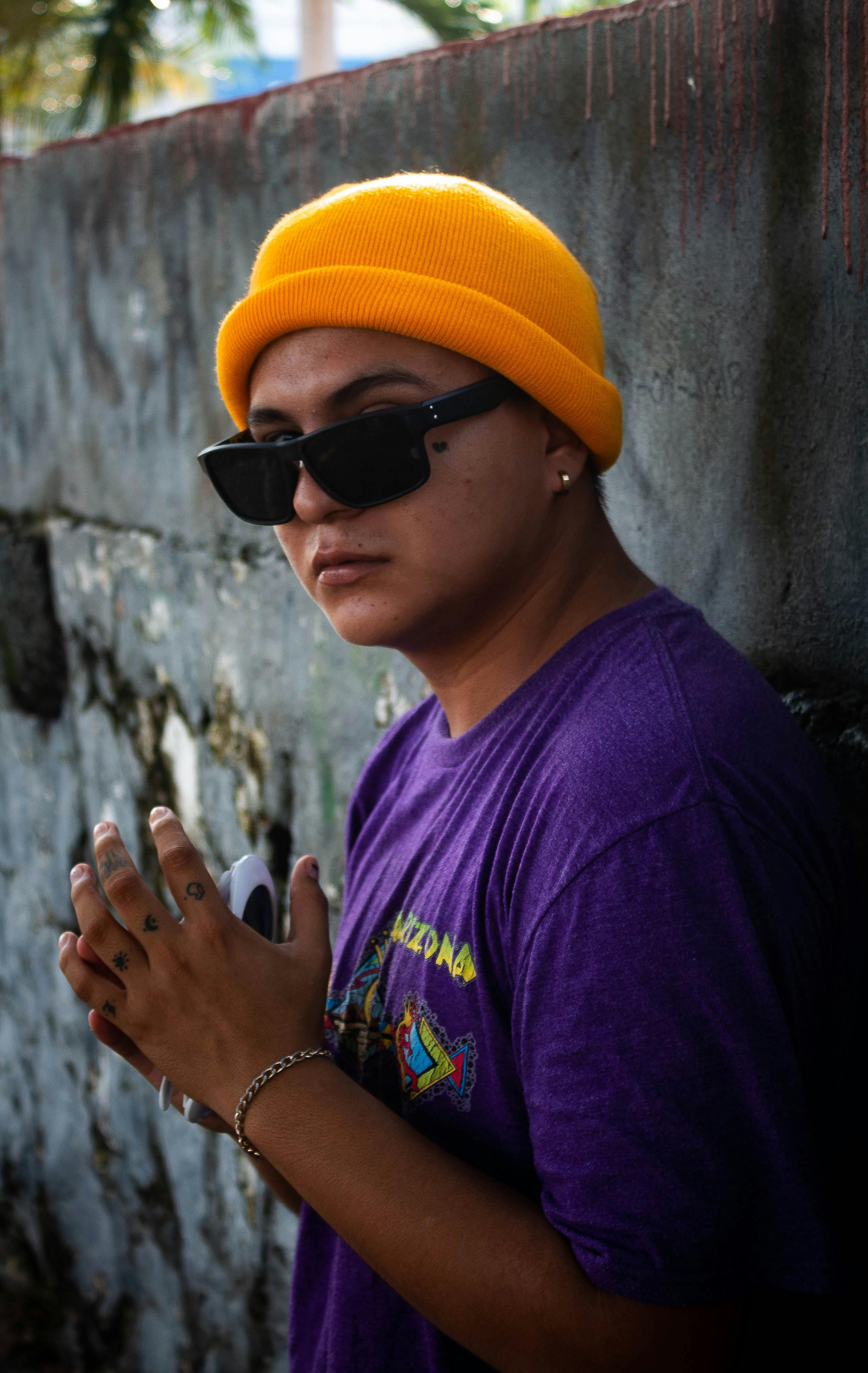 Man in a purple shirt and yellow beanie stands confidently against a rustic wall.