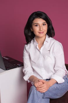 A poised woman in casual attire sits next to a laptop against a vibrant pink backdrop.