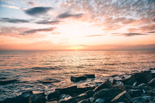 Spectacular view of coast with stones against rippled ocean under shiny cloudy sky at sundown