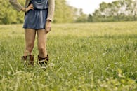 白天，一位穿着靴子和连衣裙的年轻女子站在郁郁葱葱的绿色田野里