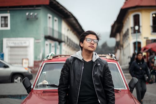 Free Stylish man in leather jacket and glasses posing in a city street, next to a red car. Stock Photo