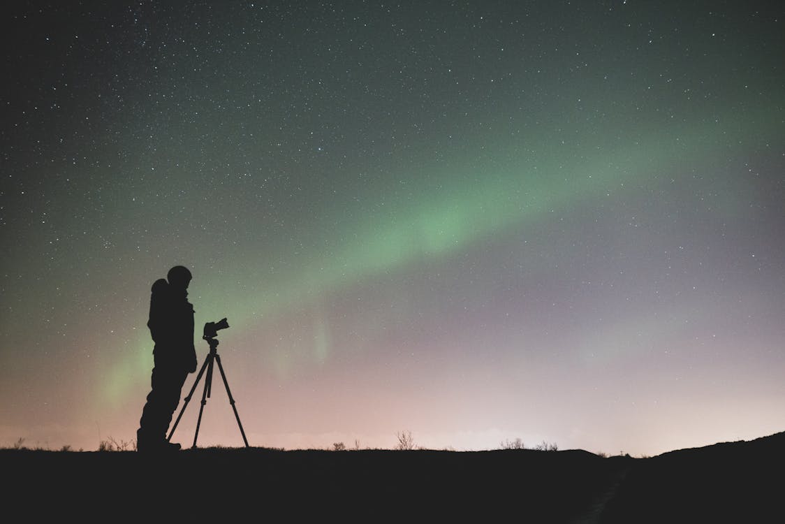 Silhouette of Person Standing Near a Tripod Under Green Sky With Stars