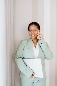 Professional woman in business attire using smartphone and holding laptop indoors.