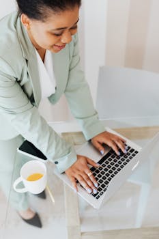 Businesswoman in a suit working on a laptop at a glass desk with coffee.