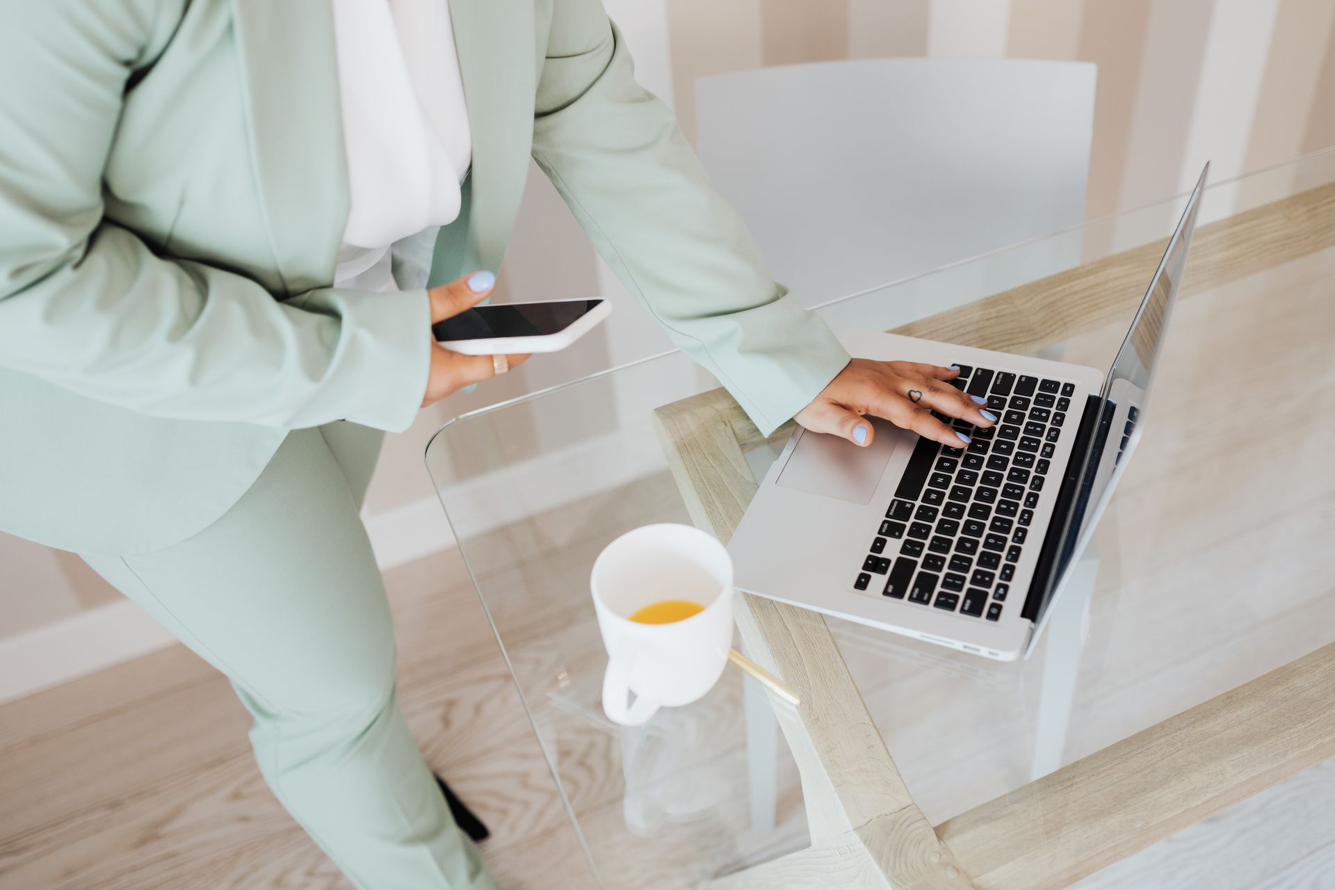 A professional using a smartphone and laptop at a sleek glass desk in an office setting.