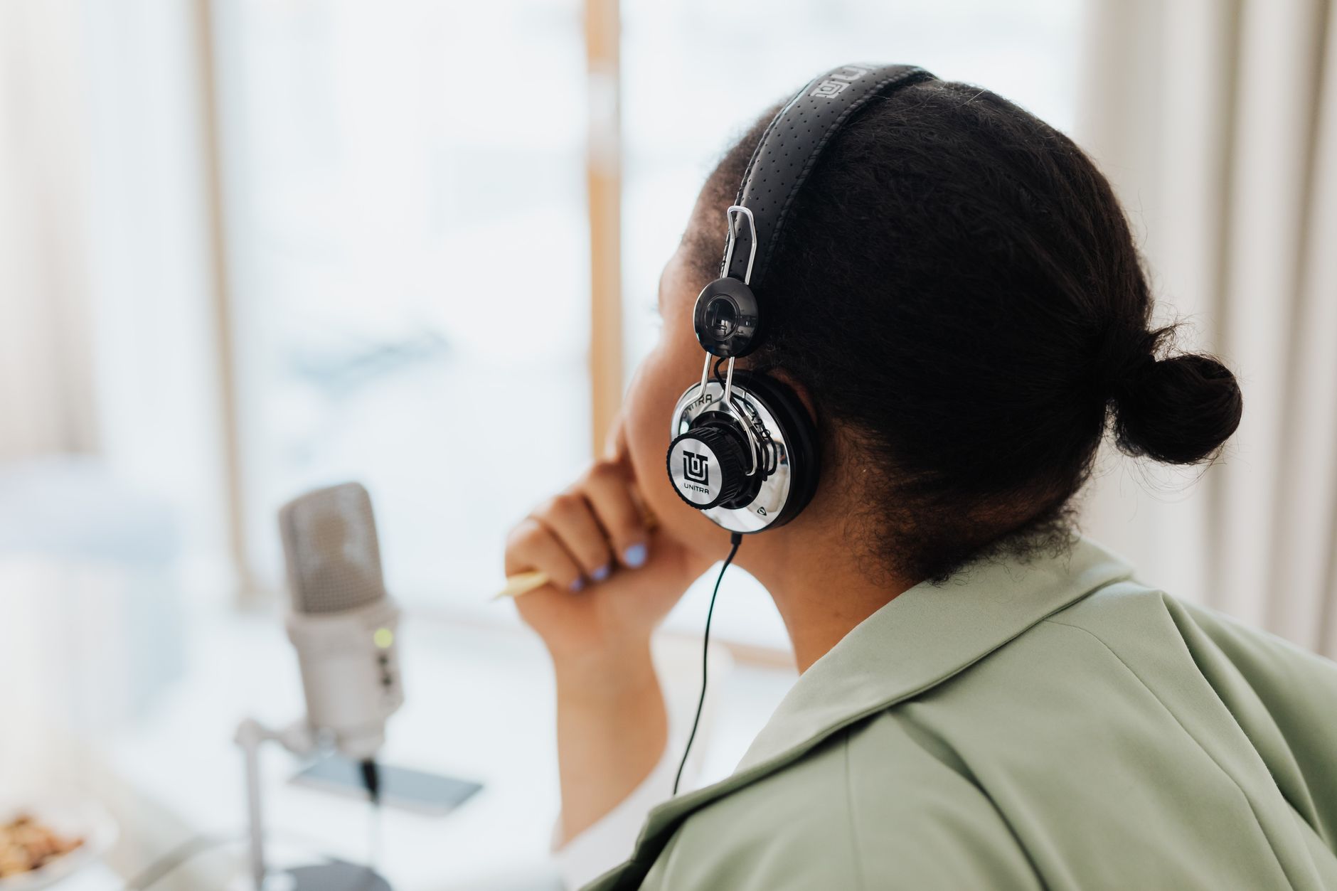 Une femme portant des écouteurs est en train de faire du podcasting à l'intérieur près d'une fenêtre.