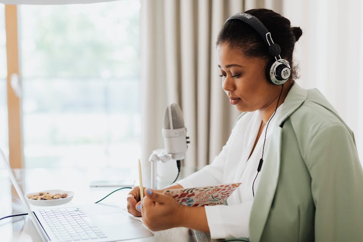 Woman Sitting In Headphones With A Laptop And A Microphone Next To Her Reading From A Notebook 