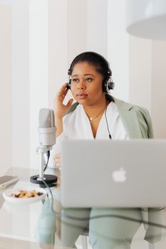 Professional woman using a laptop and microphone for podcasting in a modern workspace.