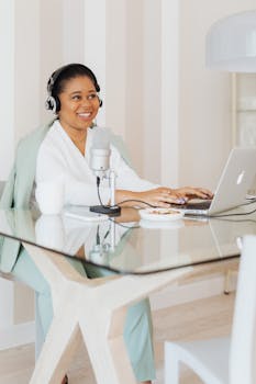 Smiling woman using laptop and microphone at glass desk in modern office setting.
