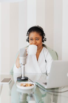 Young woman with headphones on, podcasting at home using a laptop and microphone, drinking coffee.