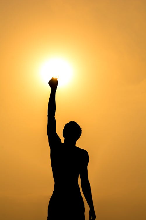 Silhouette of faceless fit male standing and raising hand against bright orange sundown sky