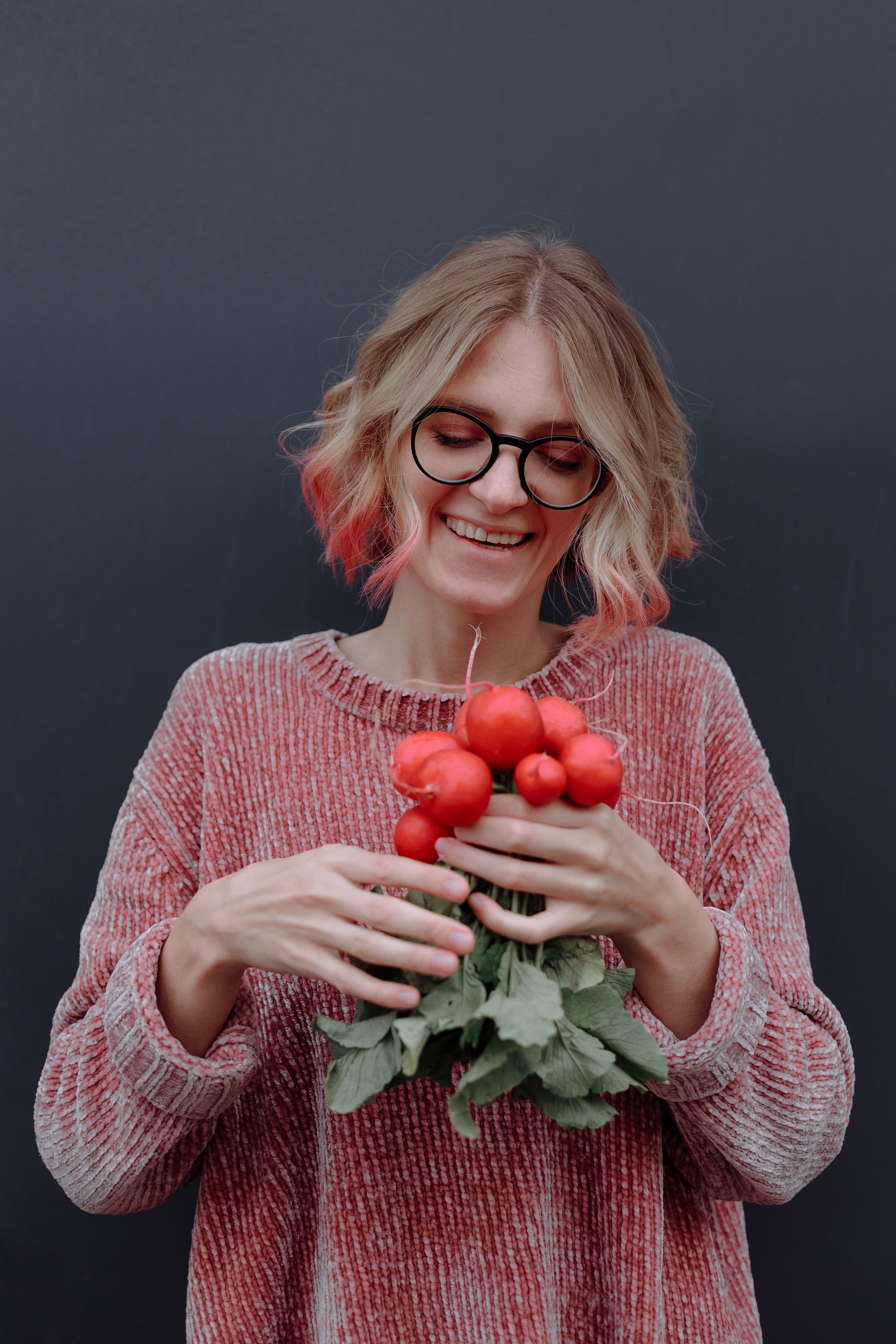 Smiling woman with glasses and red sweater holding fresh radishes, portrait style.