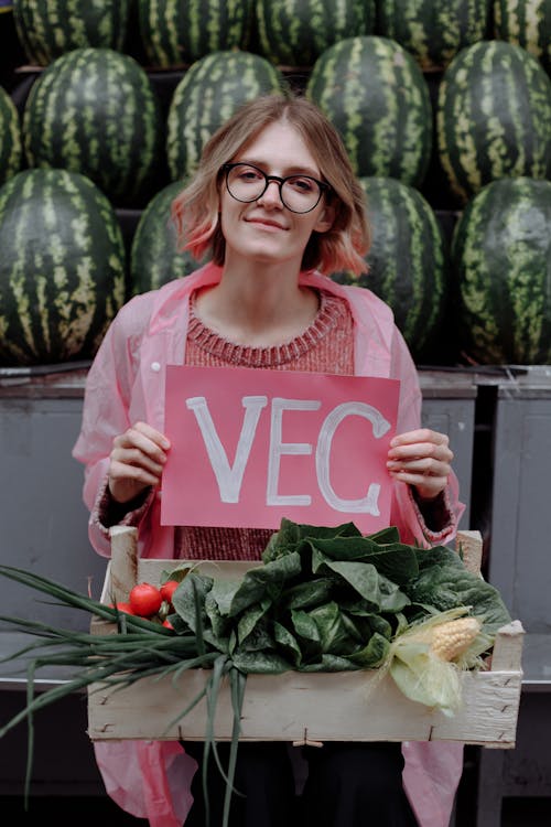 A Woman Holding a Signboard while Carrying Vegetables in a Wooden Crate