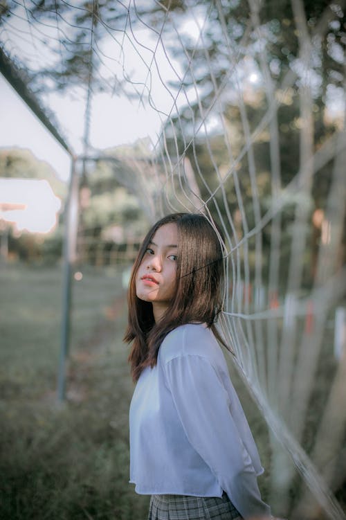 Free Pretty Asian teen girl in casual clothes with dark hair looking at camera while standing near gates with grid on blurred background Stock Photo