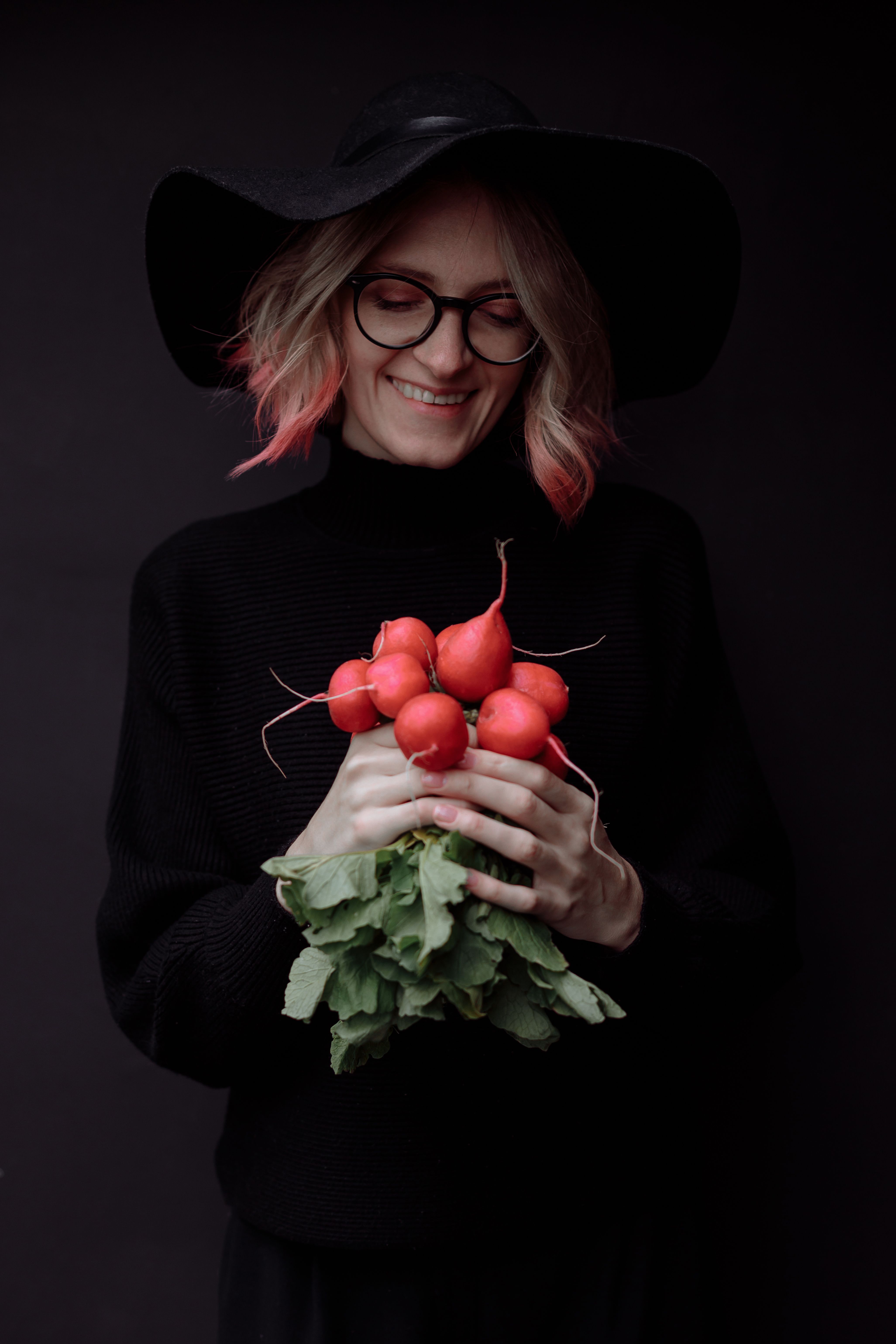 Smiling woman in a black hat holding a bunch of fresh radishes against a dark background.