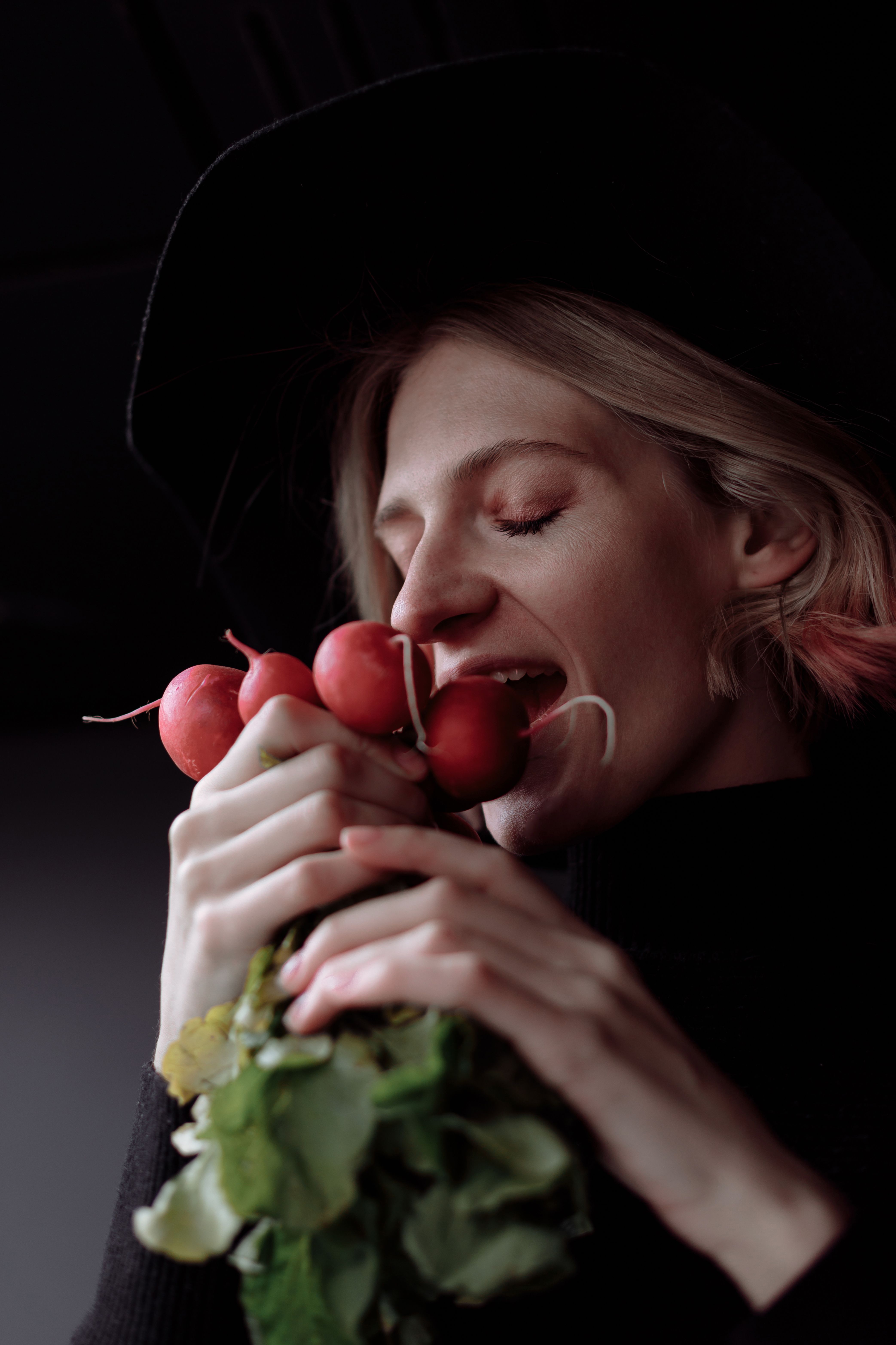 A woman in a black hat enjoys fresh radishes in a dark, moody setting.