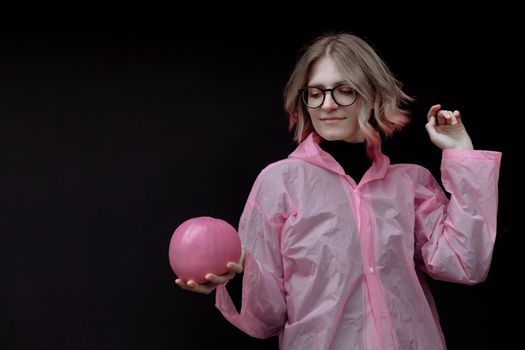 一位戴着眼镜、金发的女人，在黑色背景下身穿粉色雨衣，手捧一个粉色南瓜