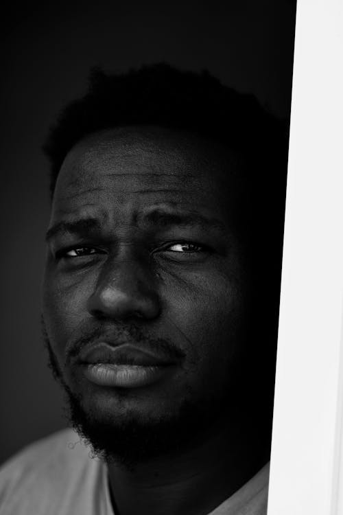 Black and white of unemotional African American male with short curly hair and beard looking at camera by drapes in dark room