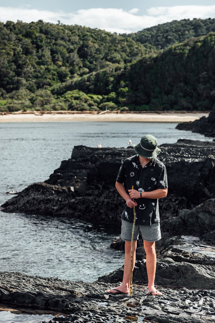 Man In Black Polo And Denim Shorts Standing On Rock Near Body Of Water