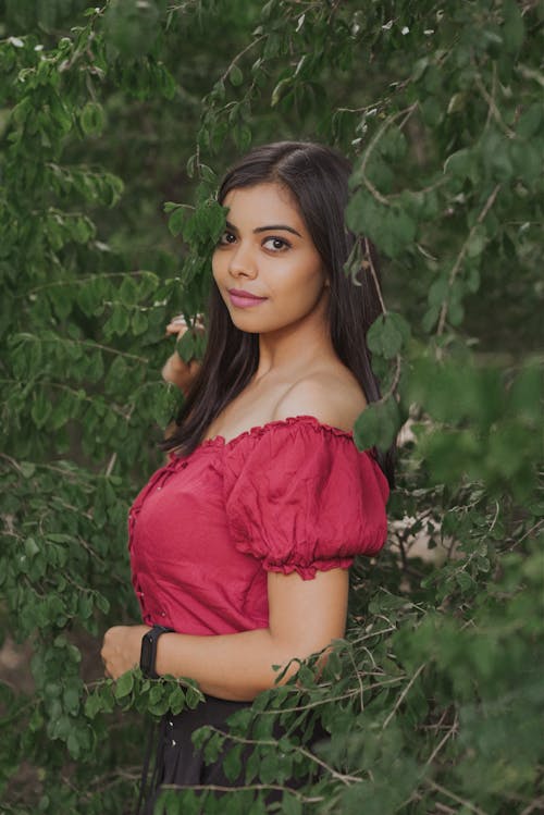 Woman in Red Off Shoulder Shirt Surrounded by Green Leaves